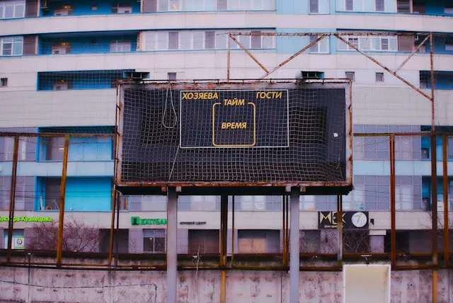 A photo of a black and white scoreboard with orange text, in Russian, on a
rusted metal frame, in a built up urban environment, with a white and blue
concrete building filling the background.
