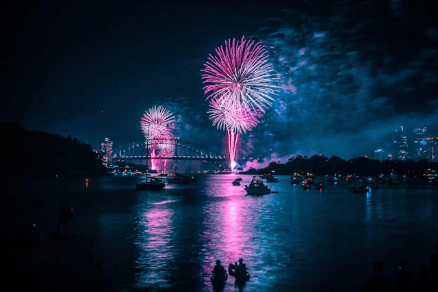 Pink fireworks explode above Syndey Harbour Bridge at nighttime, celebrating
New Years Eve, in the background, with the harbour in the foreground, filled
with boats watching the firework display.
