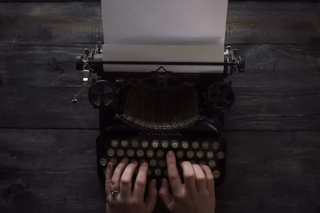 An old Corona typewriter on a wooden table, with a pair of hands resting
on the keys about to type.
