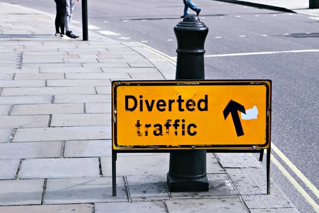A slightly worn &#039;Diverted Traffic' road sign leaning against a black bollard
on the pavement beside the road.
